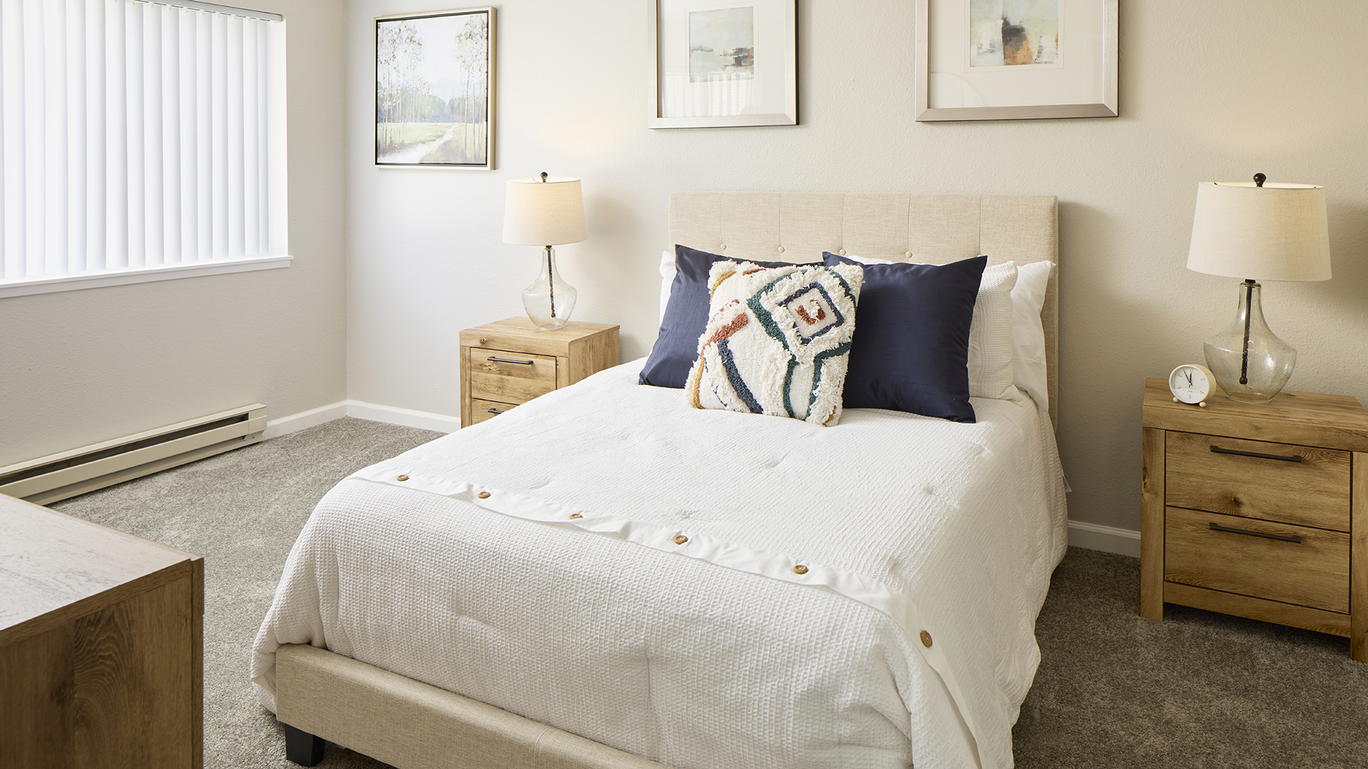 A neatly arranged Holiday Bellevue Garden apartment bedroom with a plush beige bed adorned with decorative pillows, flanked by wooden nightstands and matching table lamps, all under soft natural light